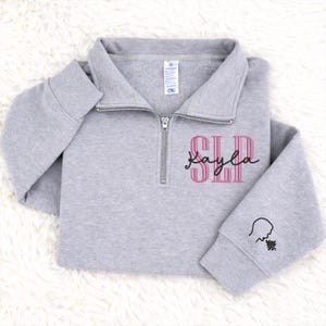 May include: A light gray quarter-zip sweatshirt with the letters "SLP" in pink and the name "Kayla" in a script font. The left sleeve has a small black graphic. The sweatshirt is made of a soft material.