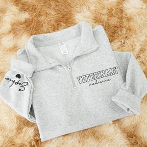 May include: A light gray quarter-zip sweatshirt with the words "VETERINARY medicine" printed in black. The left sleeve cuff has the name "Sophia" and a heart graphic. The sweatshirt is made of a soft material.