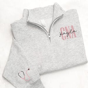 May include: A light gray quarter-zip sweatshirt with the text "CNA Kayla" in pink. A pink and black stethoscope is embroidered on the left sleeve. The sweatshirt has a silver zipper and a white tag.