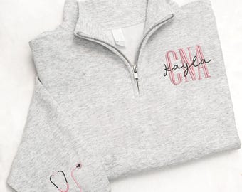 May include: A light gray quarter-zip sweatshirt with the text "CNA Kayla" in pink. A pink and black stethoscope is embroidered on the left sleeve. The sweatshirt has a silver zipper and a white tag.