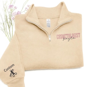 May include: A beige quarter-zip sweatshirt with the text "COSMETOLOGIST Kayla" in pink script. The left sleeve features the word "Custom" and a scissors graphic. The sweatshirt has a high collar and a silver zipper.