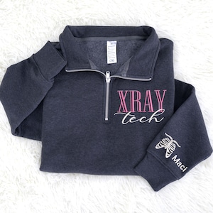 May include: Dark gray X-Ray Tech pullover sweatshirt with a quarter-zip collar. The text "XRAY tech" is embroidered in pink and white on the front. The sleeve features a skeleton graphic and the name "Maci".