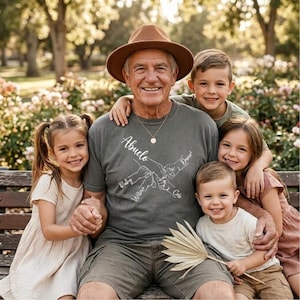 May include: A group portrait of a smiling older man wearing a brown hat and a gray t-shirt with the word "Abuelo" and names printed on it. He is surrounded by four children, all smiling. The background is a park with trees and flowers.