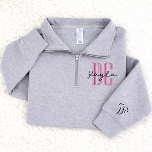 May include: A light gray pullover sweatshirt with a quarter-zip neckline. The sweatshirt features the initials "DC" in pink with the name "Kayla" in cursive script. A small design is on the sleeve.