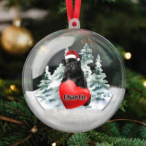 May include: A clear Christmas ornament with a black pug wearing a Santa hat, holding a red heart with the name "Charlie" on it. The ornament has a snowy base with evergreen trees in the background and a red ribbon for hanging.