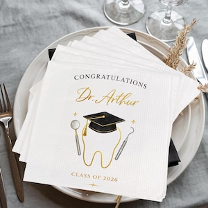 May include: White paper napkins with gold text and illustrations. The napkins read "CONGRATULATIONS Dr. Arthur" with a graduation cap, tooth, and dental tools. The text "CLASS OF 2026" is also printed on the napkins. The napkins are stacked on a white plate.