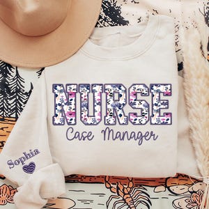 May include: Cream-colored sweatshirt with the word "NURSE" in large, floral-filled letters. Below, the text "Case Manager" is written in a cursive font. The left sleeve has the name "Sophia" embroidered with a purple heart.