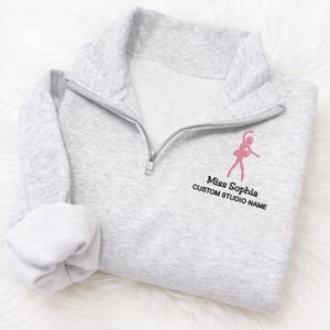 May include: A light gray quarter-zip sweatshirt with a pink embroidered ballerina design. The text "Miss Sophia" and "CUSTOM STUDIO NAME" are below the ballerina. The sweatshirt is folded with the sleeve rolled up.