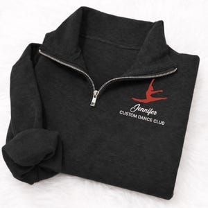 May include: Dark gray quarter-zip sweatshirt with a silver zipper. The sweatshirt features a red dancer graphic and the text "Jennifer Custom Dance Club" in white script.