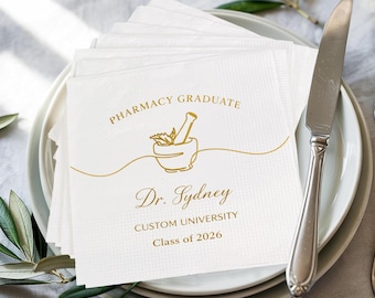 Könnte beinhalten: Ein Stapel weißer Papierservietten mit goldenem Text und einem Mörser- und Stößel-Design. Der Text lautet "Pharmacy Graduate", "Dr. Sydney", "Custom University" und "Class of 2026". Die Servietten liegen auf einem weißen Teller mit einem silbernen Messer.