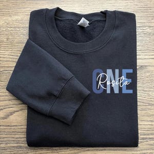 May include: Black sweatshirt with a blue and white graphic that says "ONE Rosita" on the front.