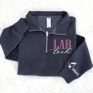 May include: Dark gray quarter-zip sweatshirt with the text "LAB tech" in pink and white script. The sleeve features a small graphic and the name "Maci". The sweatshirt has a collared neckline and a silver zipper.