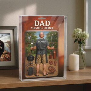 May include: A framed image featuring a man in a grilling scene, labeled "DAD THE GRILL MASTER" with the name "JAMES" on his shirt. The image includes grilling tools, food, and a small grill.