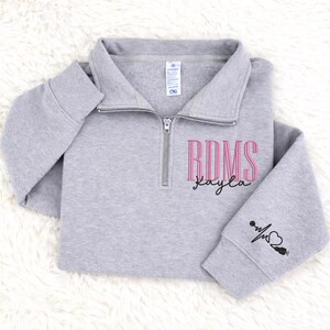 May include: A light gray quarter-zip sweatshirt with the letters "RDMS" in pink, and the name "Kayla" in a cursive font. The left sleeve has a small graphic of a heartbeat and a stethoscope. The sweatshirt is made of a soft material.