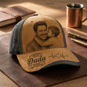 May include: A tan and charcoal gray baseball cap featuring a laser-engraved portrait of an adult and a child embracing. The front panel displays the text "BEST DADA EVER" in a decorative font alongside a heartbeat line graphic with a heart symbol.