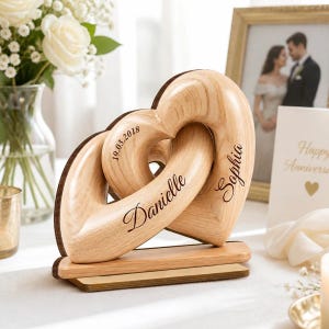 May include: A light-colored wooden sculpture featuring two interlocking heart shapes mounted on a rectangular base. The wood grain is visible throughout the piece. The names Danielle and Sophia are engraved in black script, with the date 19.03.2018.