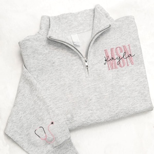 May include: A light gray quarter-zip sweatshirt with a silver zipper. The sweatshirt features the embroidered text "MSN Kayla" in pink and black, along with a pink stethoscope graphic on the sleeve. The fabric has a textured appearance.