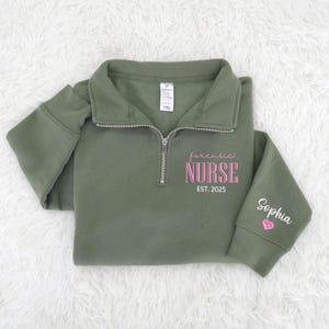 May include: An olive green quarter-zip sweatshirt with the words "forensic NURSE EST. 2025" in pink script and block letters. The sleeve has the name "Sophia" embroidered in white with a pink heart.