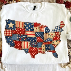 May include: White sweatshirt with a patchwork design of the United States map. The map is made of red, white, and blue fabric patches with stars, stripes, and floral patterns. A large white star is on the upper left corner of the map.