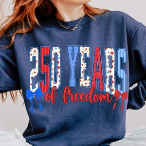 May include: Navy blue sweatshirt featuring the text "250 YEARS of freedom" in red, white, and blue. The numbers and letters have floral and polka dot patterns. Red and blue bows are at the bottom of the design.