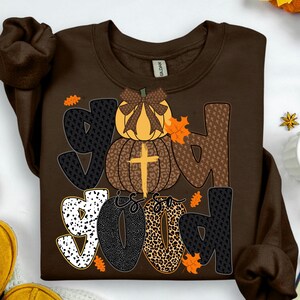 May include: A brown sweatshirt with the words "God is Good" in a fall-themed design. The word "God" is spelled out with a pumpkin and cross graphic. The word "Good" is in a mix of patterns and colors.