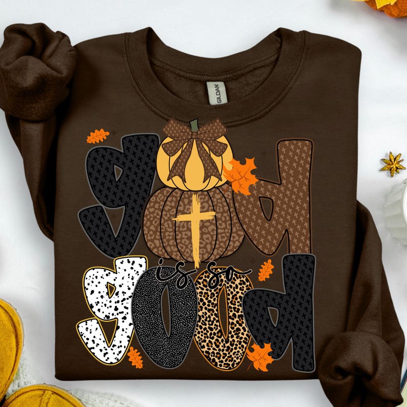 May include: A brown sweatshirt with the words "God is Good" in a fall-themed design. The word "God" is spelled out with a pumpkin and cross graphic. The word "Good" is in a mix of patterns and colors.