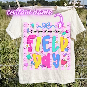 Puede incluir: Camiseta color crema con un gráfico colorido de "FIELD DAY". El diseño incluye estrellas, flores e ilustraciones escolares. El texto "Custom elementary" está encima del texto principal, y "custom name" en la parte superior.