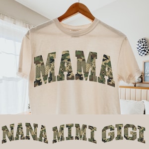 Può includere: T-shirt color crema con la scritta "MAMA" in grandi lettere con motivo mimetico. Sotto, le parole "NANA MIMI GIGI" sono visualizzate in uno stile simile. La maglietta è appesa a una gruccia di legno.