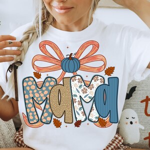 May include: White t-shirt with the word "MAMA" in a fall-themed design. The letters are decorated with pumpkins, floral patterns, and a blue bow with a pumpkin in the center. Autumn leaves are scattered around the letters.