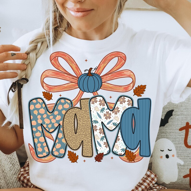 May include: White t-shirt with the word "MAMA" in a fall-themed design. The letters are decorated with pumpkins, floral patterns, and a blue bow with a pumpkin in the center. Autumn leaves are scattered around the letters.
