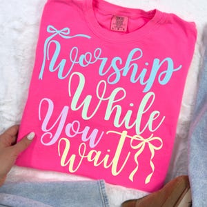 May include: A bright pink t-shirt with the words "Worship While You Wait" in pastel colors. The text is in a cursive font with decorative bows. The shirt is a Comfort Colors brand.