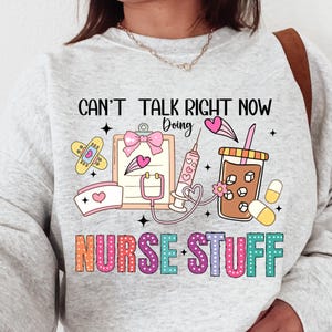 Puede incluir: Una sudadera gris claro con el texto "CAN'T TALK RIGHT NOW Doing" y "NURSE STUFF" en letras coloridas. El diseño incluye ilustraciones de temática médica como una carpeta, una jeringa y una taza de café.