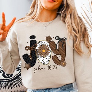 May include: Beige sweatshirt with the word "JOY" in a decorative font. The "J" and "Y" are brown with a floral pattern, and the "O" is white with a brown flower. The text "john 16:22" is printed below the word "JOY".