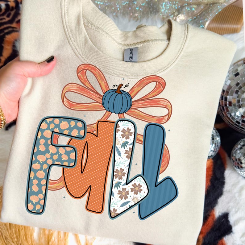 May include: Cream-colored sweatshirt with the word "FALL" in a colorful design. The "F" is patterned with small pumpkins, the "A" is orange with dots, and the "LL" is floral and striped. A blue pumpkin and orange bow complete the design.