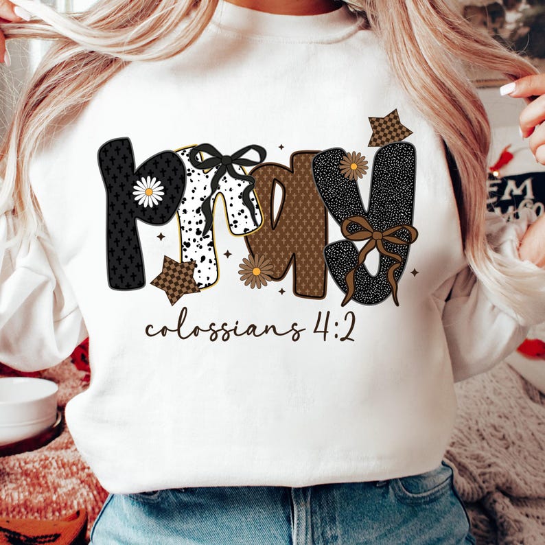 May include: White sweatshirt with the word "PRAY" in a decorative font. The letters are black, white, and brown with floral and patterned accents. The text "colossians 4:2" is below the word "PRAY".