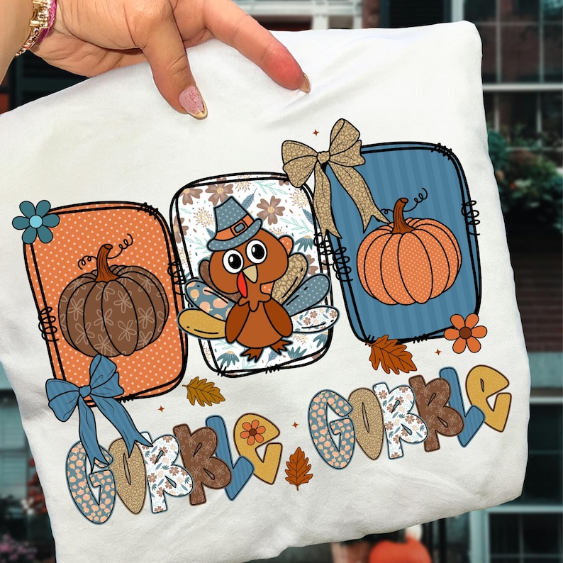 May include: White t-shirt featuring a Thanksgiving-themed design. The design includes a cartoon turkey wearing a pilgrim hat, pumpkins, and the words "Gobble Gobble" in a decorative font. The design is framed by decorative borders and bows.