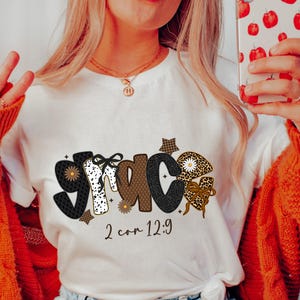 May include: White t-shirt with the word "GRACE" in a decorative font. The letters are adorned with patterns like polka dots, leopard print, and gingham. The text "2 cor 12:9" is printed below the word.