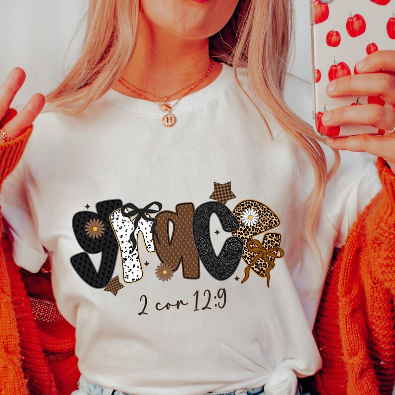 May include: White t-shirt with the word "GRACE" in a decorative font. The letters are adorned with patterns like polka dots, leopard print, and gingham. The text "2 cor 12:9" is printed below the word.