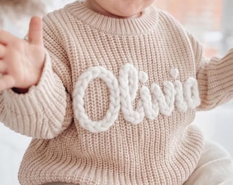 Könnte beinhalten: Ein beigefarbener Strickpullover mit dem Namen Olivia in großen, weißen, geflochtenen Garnbuchstaben auf der Brust. Der Pullover hat einen gerippten Rundhalsausschnitt und gerippte Bündchen. Ein passendes beiges Stoffstirnband mit Schleife.