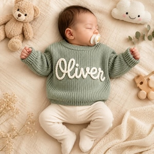 May include: A sleeping infant wearing a sage green long-sleeve knit sweater with the name Oliver embroidered in white cursive script across the chest, paired with cream-colored ribbed leggings and a white pacifier in their mouth.