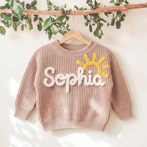May include: A light pink knit sweater with the name "Sophia" in white script and a yellow sun design. The sweater is hanging on a wooden hanger against a white background with greenery.
