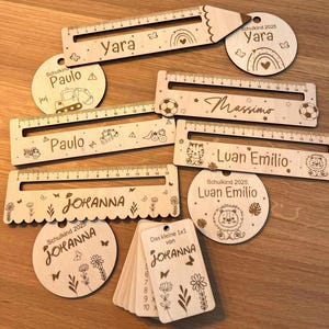 Könnte beinhalten: Holzlineale und runde Etiketten mit eingravierten Namen und Designs. Die Lineale messen bis zu 35,5 cm und tragen Namen wie Yara, Paulo, Massimo, Johanna und Luan Emilio. Die Etiketten enthalten Illustrationen von einem Regenbogen, Fussbällen und einem Löwen.