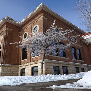 May include: A brick building with a snow-covered roof and a tree in front. The building has a large arched entrance and several windows. The tree is bare and covered in frost.