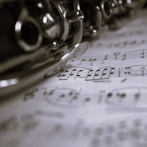 May include: A silver clarinet rests on a sheet of music with musical notes printed on it.