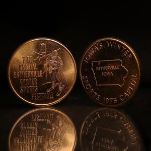 May include: Two bronze-colored commemorative coins. The first coin features a skier and the text "7th Annual Estherville Winter Sports Festival". The second coin features the text "Iowa's Winter Sports Capital 1975" and an outline of the state of Iowa with a star and the text "Estherville Iowa".