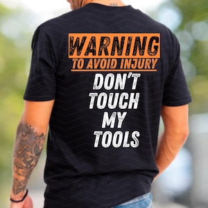 Puede incluir: Camiseta negra con el texto "WARNING TO AVOID INJURY DON'T TOUCH MY TOOLS" en naranja y blanco.