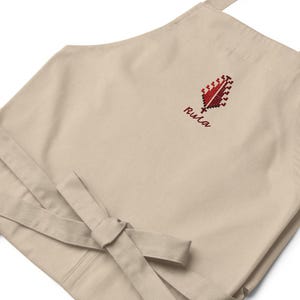 Peut inclure: Tablier beige avec un motif de plume brodée rouge et noir et le mot "Rula" en écriture rouge. Le tablier a une taille nouée et une sangle de cou. Le tablier est fait d'un tissu doux et durable.