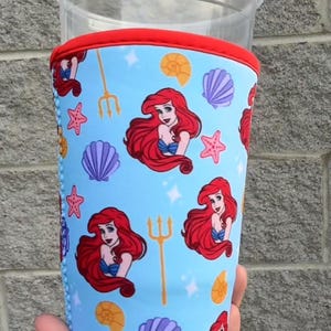 Può includere: Una custodia blu per bevande con un motivo ripetuto di una sirena dai capelli rossi, conchiglie, stelle marine e tridenti. I bordi superiore e inferiore sono rifiniti in rosso e blu. La custodia è progettata per adattarsi a un bicchiere di plastica trasparente.