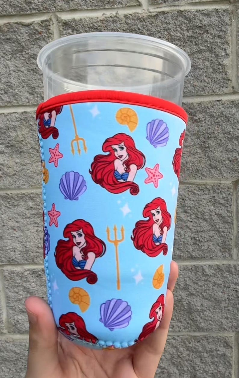 May include: A blue drink sleeve featuring a repeating pattern of a red-haired mermaid, seashells, starfish, and tridents. The top and bottom edges are trimmed in red and blue. The sleeve is designed to fit a clear plastic cup.