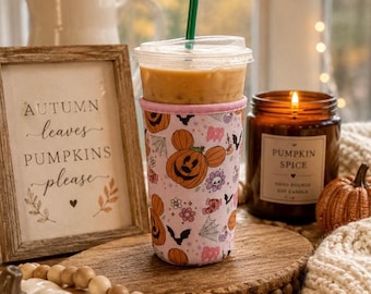 Könnte beinhalten: Ein durchsichtiger Eiskaffeebecher aus Kunststoff mit grünem Strohhalm, umhüllt von einer hellrosa Stoffhülle mit einem Muster aus orangefarbenen Kürbissen in Mickey-Mouse-Form, schwarzen Fledermäusen, weißen Spinnennetzen und dem Wort BOO.